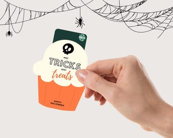 May include: A Halloween-themed gift card shaped like a cupcake, held by a hand. The cupcake is orange with white frosting and features the text "Not Tricks Just Treats" and a skull. A spider web and spiders are in the background.