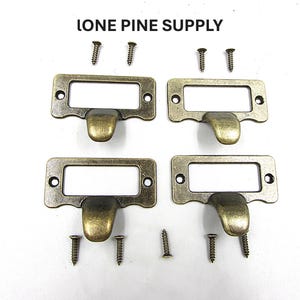 May include: Four antique brass label holders with screws. Each holder has a rectangular frame with a curved handle below. The label holders and screws are on a white background. The text "IONE PINE SUPPLY" is at the top.