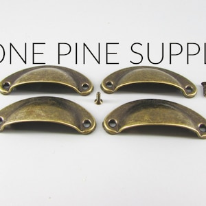 May include: Four antique brass-colored cup pull handles with matching screws. The handles are in a half-moon shape with two holes for screws. The text "LONE PINE SUPPLY" is printed above the handles.