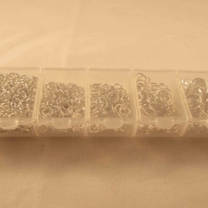 May include: A clear plastic storage box with seven compartments filled with silver jump rings. The jump rings are of various sizes.