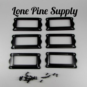 May include: Six black metal label holders with rounded edges and screw holes. The label holders are arranged in two rows of three. A pile of black screws is below the label holders.