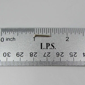 May include: A close-up of a silver metal pin with a bent end, measured against a ruler. The pin is approximately 1 inch long. The ruler reads "L.P.S."