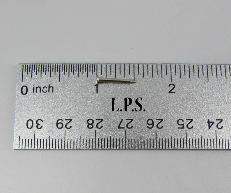 May include: A close-up of a silver metal pin with a bent end, measured against a ruler. The pin is approximately 1 inch long. The ruler reads "L.P.S."