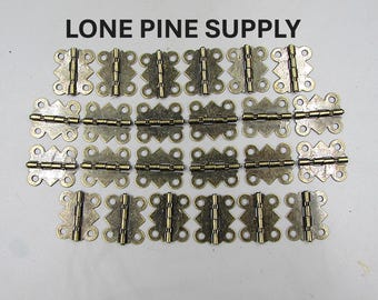 Könnte beinhalten: Eine Sammlung von antikfarbenen Messing-Scharnieren, jedes mit einem dekorativen, verzierten Design. Die Scharniere sind auf einer hellen Oberfläche angeordnet. Der Text "LONE PINE SUPPLY" ist oben im Bild aufgedruckt.