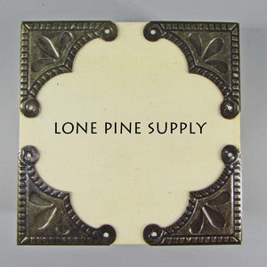 May include: A square, off-white wooden plaque with the text "LONE PINE SUPPLY" in black. The corners are adorned with decorative, antique brass-colored metal corner brackets, each featuring a floral design.
