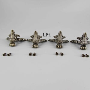 May include: Four antique gold-toned metal furniture corner brackets with ornate designs. Each bracket comes with two screws for installation. L.P.S. is written in the center of the image.