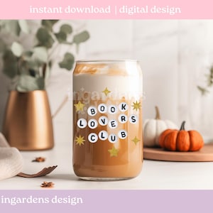 Pu&ograve; includere: Un bicchiere trasparente a forma di lattina pieno di una bevanda a strati. Il bicchiere presenta la scritta "BOOK LOVERS CLUB" in un design circolare con stelle dorate. L'immagine include anche zucche e un vaso dorato.