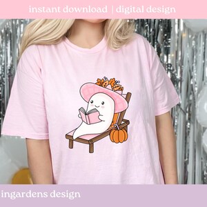 May include: A light pink t-shirt featuring a cartoon ghost reading a book while relaxing on a beach chair. The ghost wears a pink hat and is surrounded by fall foliage and a pumpkin. Text at the top reads "instant download | digital design."