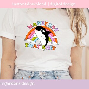 K&ouml;nnte beinhalten: Wei&szlig;es T-Shirt mit einem Cartoon-Orca-Wal-Design, einem Regenbogen und dem Text "MANIFEST THAT SHIT". Das Design verwendet helle Farben. Das T-Shirt hat einen l&auml;ssigen Stil.