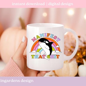 K&ouml;nnte beinhalten: Wei&szlig;e Keramik-Tasse mit einem Cartoon-Orca-Wal-Design, Regenbogen und dem Text "MANIFEST THAT SHIT". Die Tasse hat einen gebogenen Griff und ist f&uuml;r hei&szlig;e oder kalte Getr&auml;nke geeignet.