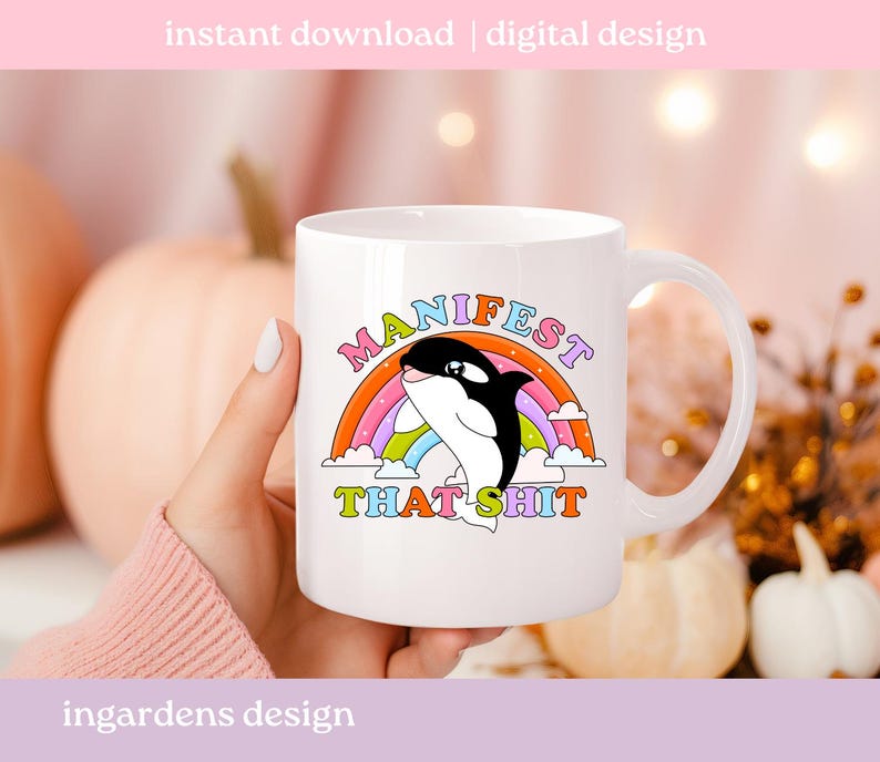 K&ouml;nnte beinhalten: Wei&szlig;e Keramik-Tasse mit einem Cartoon-Orca-Wal-Design, Regenbogen und dem Text "MANIFEST THAT SHIT". Die Tasse hat einen gebogenen Griff und ist f&uuml;r hei&szlig;e oder kalte Getr&auml;nke geeignet.