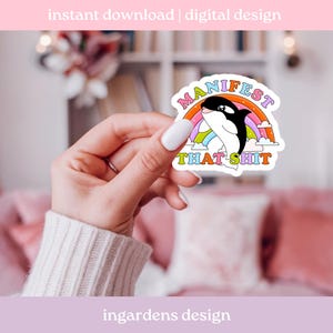 K&ouml;nnte beinhalten: Ein Aufkleber mit einem Cartoon-Orca mit einem Regenbogen und dem Text "MANIFEST THAT SHIT". Der Orca ist schwarz und wei&szlig;, und der Regenbogen hat mehrere Farben. Der Aufkleber wird von einer Hand gehalten.