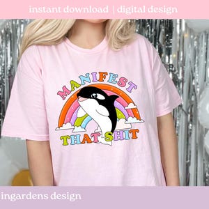 K&ouml;nnte beinhalten: Hellrosa T-Shirt mit einer Grafik eines schwarz-wei&szlig;en Orcas, der durch einen Regenbogen springt. Der Text "MANIFEST THAT SHIT" befindet sich &uuml;ber und unter dem Orca. Die Worte "sofortiger Download | digitales Design" stehen oben.