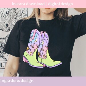 May include: A black t-shirt featuring a graphic of two colourful cowboy boots with a celestial design. The boots are green, pink and blue with stars, moons and planets.