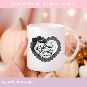 May include: White ceramic mug featuring a heart-shaped design with the text "The Shadow Daddy book club". The design includes a black bow and a gray scalloped border. The words "instant download | digital design" and "ingardens design" are also visible.
