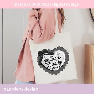 May include: A white tote bag featuring a black and gray heart-shaped design with a bow and the text "the Shadow Daddy book club." The bag has a simple design with the words "instant download | digital design" and "ingardens design" printed on it.