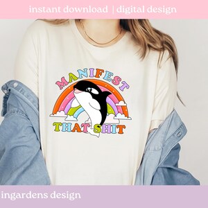 K&ouml;nnte beinhalten: Cremefarbenes T-Shirt mit einer Grafik eines schwarz-wei&szlig;en Orcas, der &uuml;ber einen Regenbogen springt. Der Text "MANIFEST THAT SHIT" ist in bunten Buchstaben &uuml;ber dem Orca angeordnet. Die Worte "sofortiger Download | digitales Design" stehen oben.