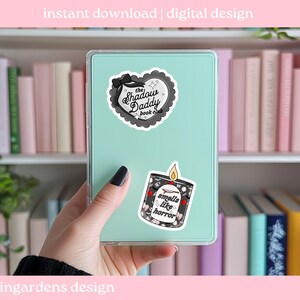 May include: A teal tablet case with two stickers: one heart-shaped with "The Shadow Daddy book club" text, and another of a candle with "smells like horror" text. The background includes a bookshelf with pink and white books.