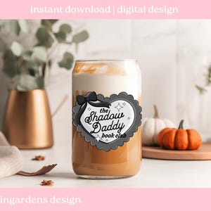 May include: A clear glass can-shaped cup filled with a layered beverage, featuring a creamy white top and a light brown base. The cup has a decorative label with the text "The Shadow Daddy Book Club". The image also includes pumpkins and decorative elements.