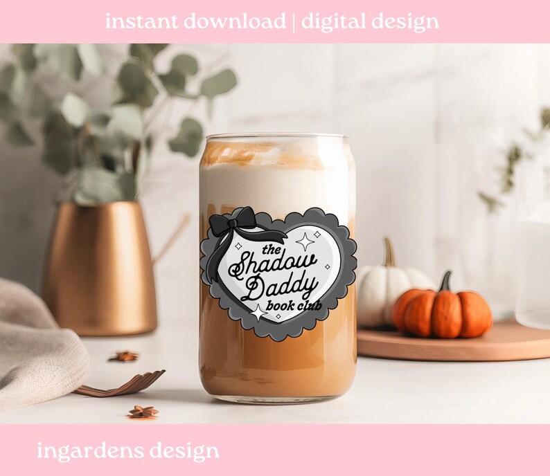 May include: A clear glass can-shaped cup filled with a layered beverage, featuring a creamy white top and a light brown base. The cup has a decorative label with the text "The Shadow Daddy Book Club". The image also includes pumpkins and decorative elements.