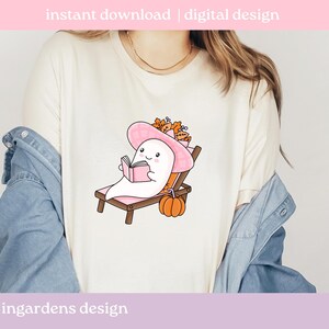 May include: White t-shirt featuring a cartoon ghost wearing a pink sun hat and reading a book while relaxing on a wooden beach chair. An orange pumpkin sits nearby. Text at the top reads "instant download | digital design" and at the bottom "ingardens design".