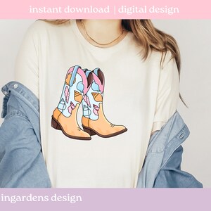 May include: A cream-colored t-shirt featuring a colorful illustration of cowboy boots. The boots are light tan with blue, pink, and white accents, and are decorated with seashells and feathers. Text at the top reads "instant download | digital design".