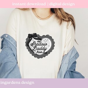 May include: Cream-colored t-shirt featuring a heart-shaped graphic with the text "the Shadow Daddy book club". The graphic is in black and gray with a bow and star accents. The words "instant download | digital design" are at the top.