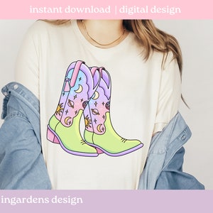 May include: A white t-shirt featuring a graphic of two colourful cowboy boots with a celestial design. The boots are decorated with stars, moons and planets. The text "ingardens design" is at the bottom of the shirt.