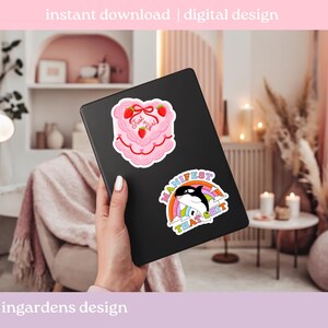 K&ouml;nnte beinhalten: Ein schwarzes Tablet mit zwei Aufklebern. Einer ist ein rosa Kuchen mit Erdbeeren und dem Text "Just a Girl". Der andere ist ein Regenbogen mit einem Wal und dem Text "Manifest That Shit".