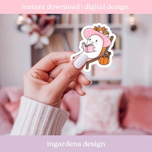 May include: A sticker featuring a cartoon ghost wearing a pink hat and reading a book while relaxing on a beach chair. The sticker has a pumpkin and fall foliage. Text at the top says "instant download | digital design".