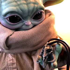 May include: A green and brown plush toy wearing a brown scarf holds a silver pendant with a skull design. The pendant is on a brown leather cord.