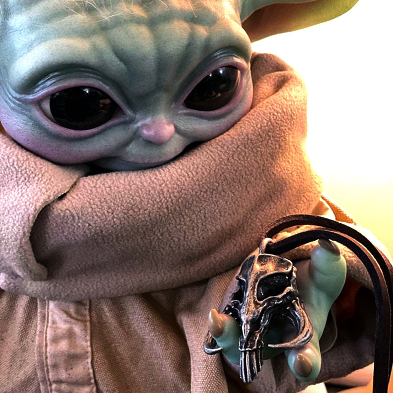 May include: A green and brown plush toy wearing a brown scarf holds a silver pendant with a skull design. The pendant is on a brown leather cord.