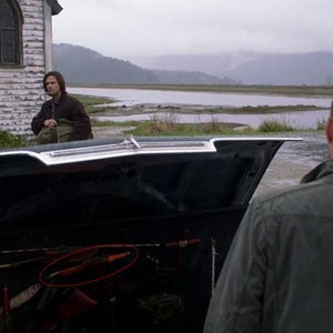 May include: The open boot of a black car contains a variety of hunting and survival gear, including a long, wooden-handled knife, a bow and arrow, and a metal axe.