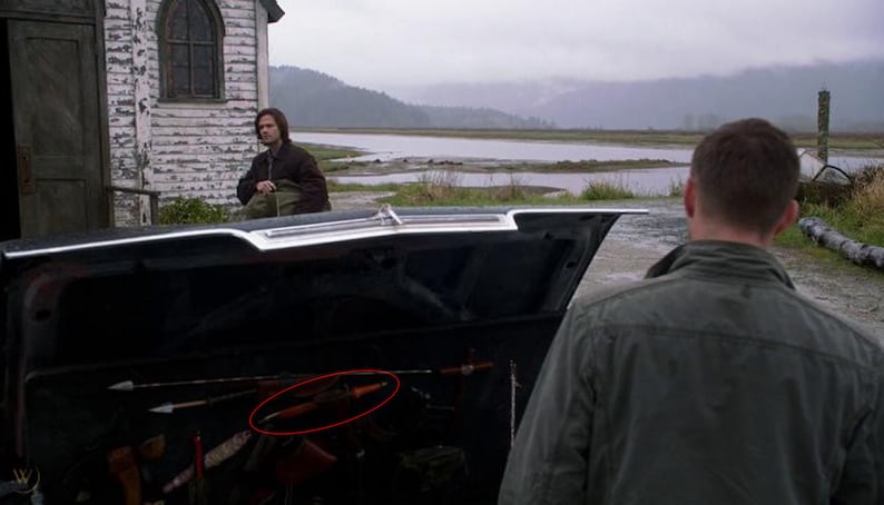 May include: The open boot of a black car contains a variety of hunting and survival gear, including a long, wooden-handled knife, a bow and arrow, and a metal axe.
