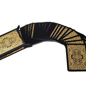 Może przedstawiać: Talia kart do gry z czarnym tłem i złotym wzorem filigranowym. Karty są rozłożone wachlarzem, ukazując skomplikowane wzory na rewersach kart. Pudełko na karty r&oacute;wnież posiada ten sam złoty wz&oacute;r.