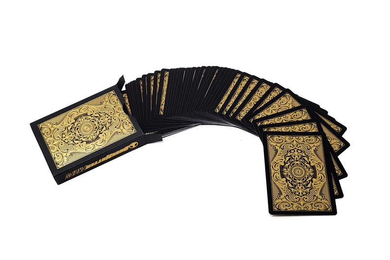 Może przedstawiać: Talia kart do gry z czarnym tłem i złotym wzorem filigranowym. Karty są rozłożone wachlarzem, ukazując skomplikowane wzory na rewersach kart. Pudełko na karty r&oacute;wnież posiada ten sam złoty wz&oacute;r.