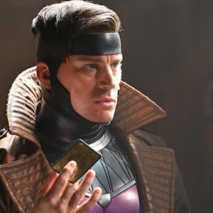 Może przedstawiać: Osoba w brązowym sk&oacute;rzanym płaszczu i czarnej opasce na głowę trzyma kartę do gry. Postać nosi czarno-fioletowy kostium z wysokim kołnierzem i brązowy sk&oacute;rzany płaszcz.