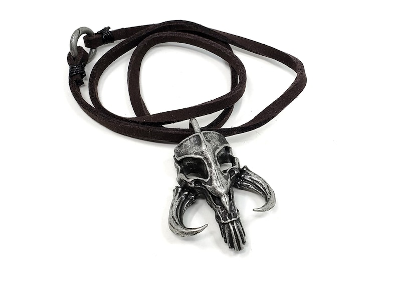 May include: A silver-toned pendant necklace featuring a detailed skull with horns. The necklace is made of brown leather cord.