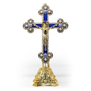 Pode incluir: Cruz dourada ornamentada com fundo de esmalte azul e detalhes dourados. A cruz apresenta uma figura dourada de Jesus Cristo e é adornada com pérolas falsas. A cruz é montada sobre uma base dourada decorativa.
