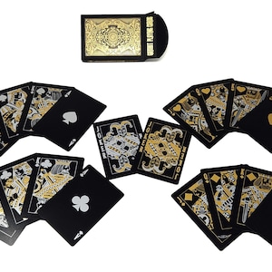 Może przedstawiać: Talia kart do gry z czarnym tłem i złotymi i srebrnymi detalami. Karty są rozłożone wachlarzem, prezentując kolory i figury. Pudełko na karty jest czarne ze złotym filigranem i napisem "COURT PLAYING CARDS".