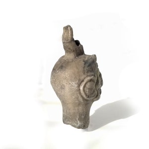 May include: A ceramic sculpture of a human head with a spout on top. The head is gray and has a textured surface. The sculpture is on a white background.