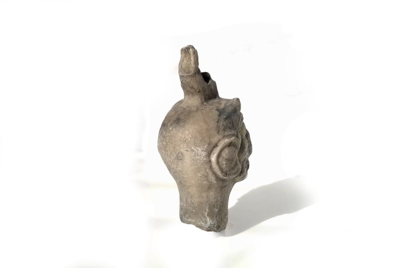 May include: A ceramic sculpture of a human head with a spout on top. The head is gray and has a textured surface. The sculpture is on a white background.