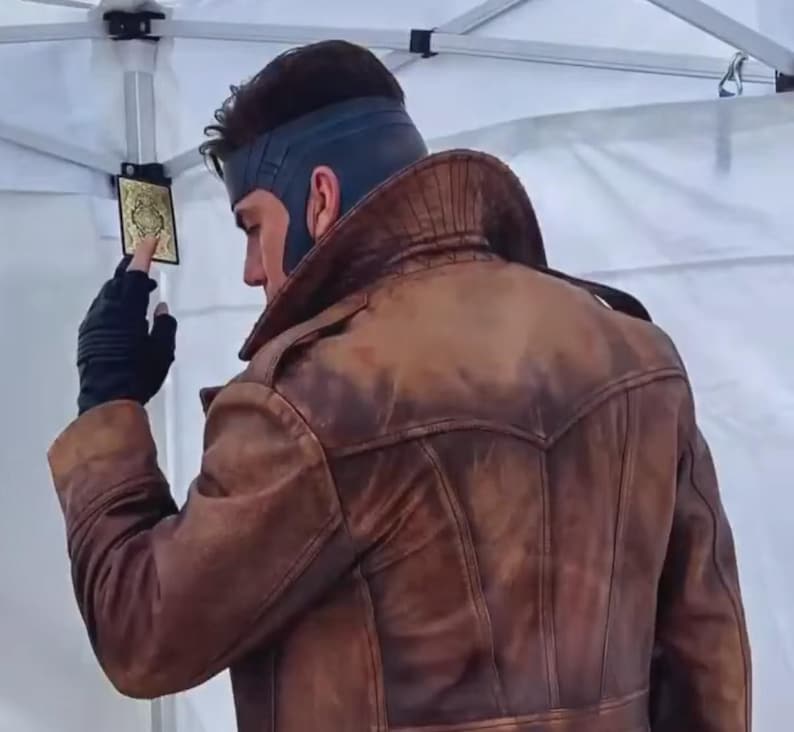 Może przedstawiać: Brązowa sk&oacute;rzana kurtka z postarzanym wykończeniem. Kurtka ma szeroki kołnierz i dopasowany kr&oacute;j. Osoba ma na sobie niebieską maskę i czarne rękawiczki, trzymając kartę do gry ze złotym wzorem.