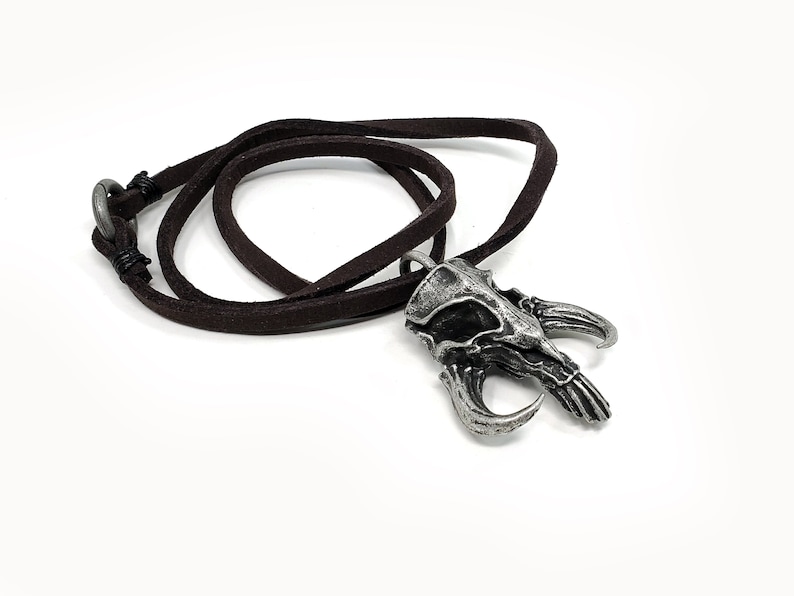 May include: A silver pendant necklace with a detailed skull design and horns. The necklace is on a brown leather cord.