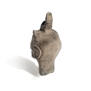 May include: A gray ceramic sculpture of a head with a pointed top. The sculpture has a detailed face with a large ear and a small mouth.