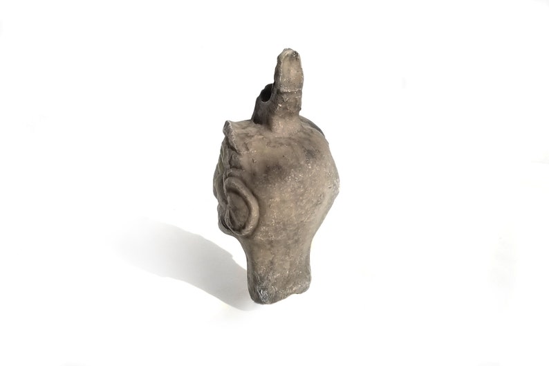 May include: A gray ceramic sculpture of a head with a pointed top. The sculpture has a detailed face with a large ear and a small mouth.