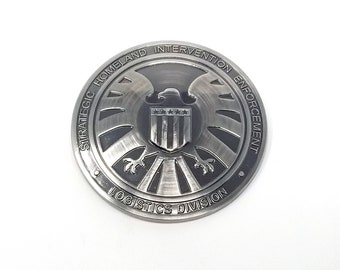 May include: A silver metal coin with a black and white image of a shield with a hawk and stars. The text around the shield reads "Strategic Homeland Intervention Enforcement Logistics Division".