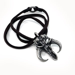 May include: A silver-toned pendant necklace with a detailed skull design and two horns. The necklace is on a brown leather cord.