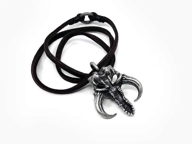 May include: A silver-toned pendant necklace with a detailed skull design and two horns. The necklace is on a brown leather cord.
