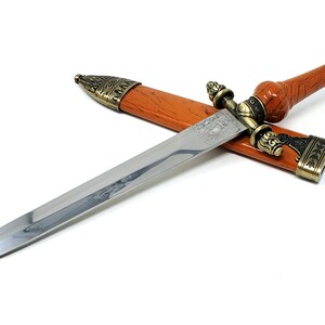 May include: A silver dagger with a brown wooden handle and ornate metal accents. The dagger is in a brown leather sheath with a metal tip.